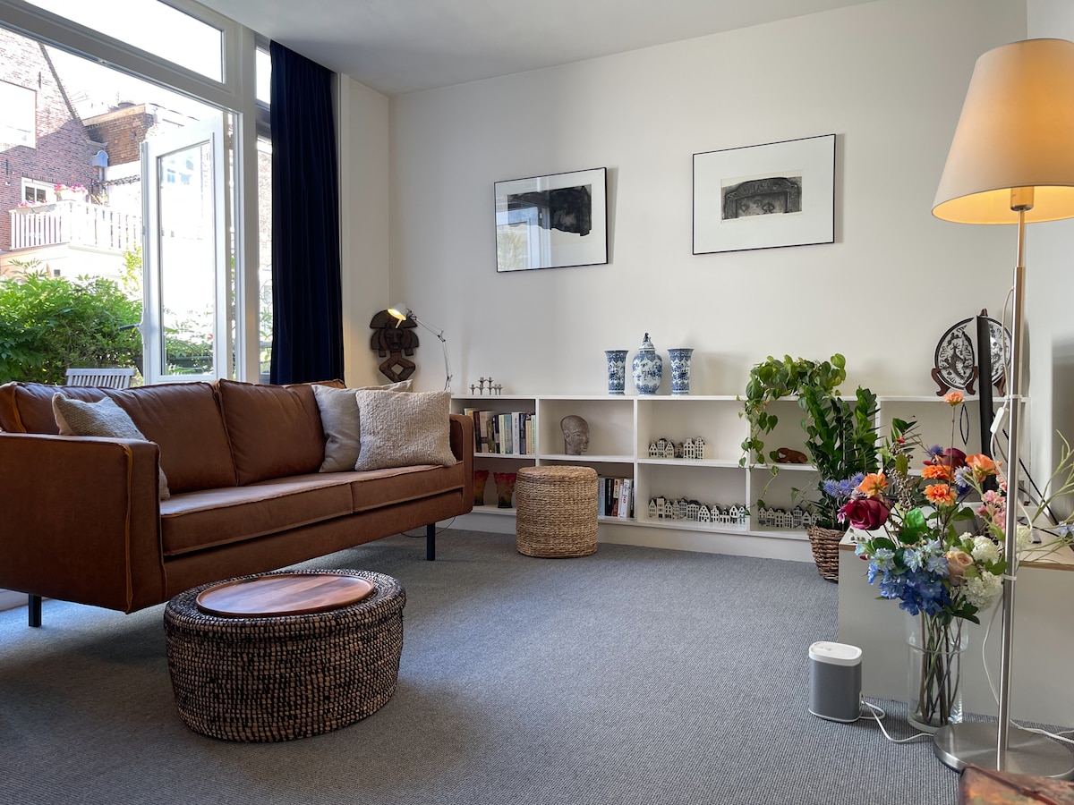 A comfortable living room is illuminated by natural light. A brown sofa, accented with cushions, is positioned in front of a low table. A shelf displays decorative items. Potted plants and flowers add a touch of color, while nearby windows offer a view of the outdoor area.