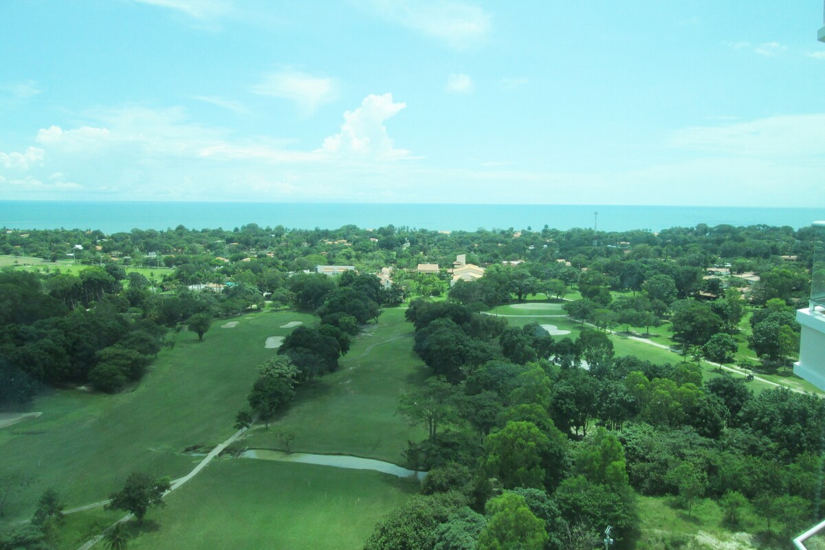 A scenic view showcases lush greenery and well-maintained golf course landscapes, stretching towards the horizon where the Pacific Ocean meets the sky. The image captures a tranquil atmosphere with a blend of trees and manicured fairways.
