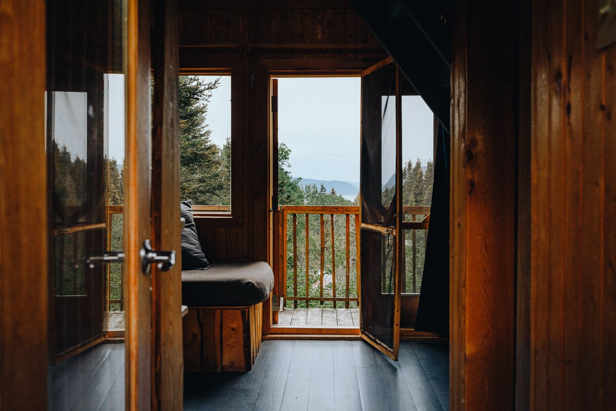 A wooden interior reveals two open doors leading to a balcony, providing a view of surrounding trees and mountains. A comfortable seating area, with a cushioned bench, is positioned near the entrance, enhancing the inviting space filled with natural light.