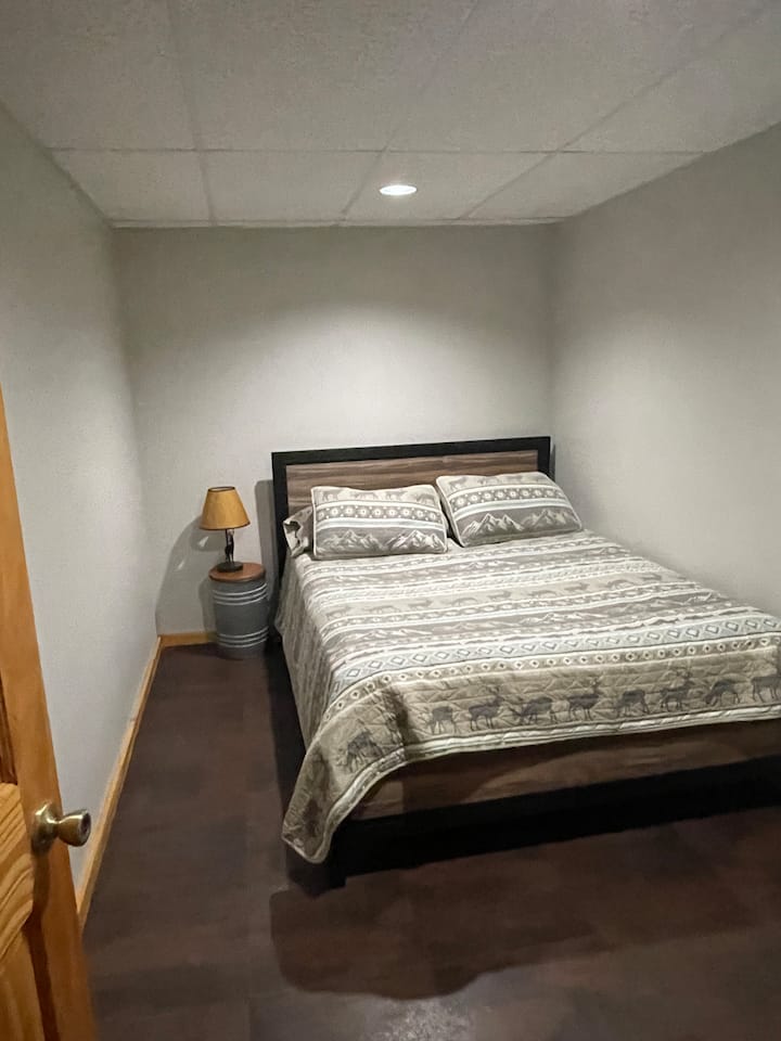 Dormitorio sótano con una cama tamaño queen ubicada junto al espacio de reunión del sótano.