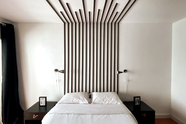 The bedroom features a spacious bed with a minimalist headboard design composed of vertical wooden slats. Nightstands on either side hold modern lamps. Natural light illuminates the room through a large window, with dark curtains providing an option for privacy.