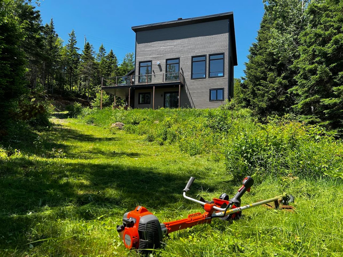A modern chalet is visible in the background, characterized by large windows and a multi-level design. In the foreground, a red grass trimmer rests on a grassy area, surrounded by lush greenery and trees, reflecting the natural landscape of the surroundings.