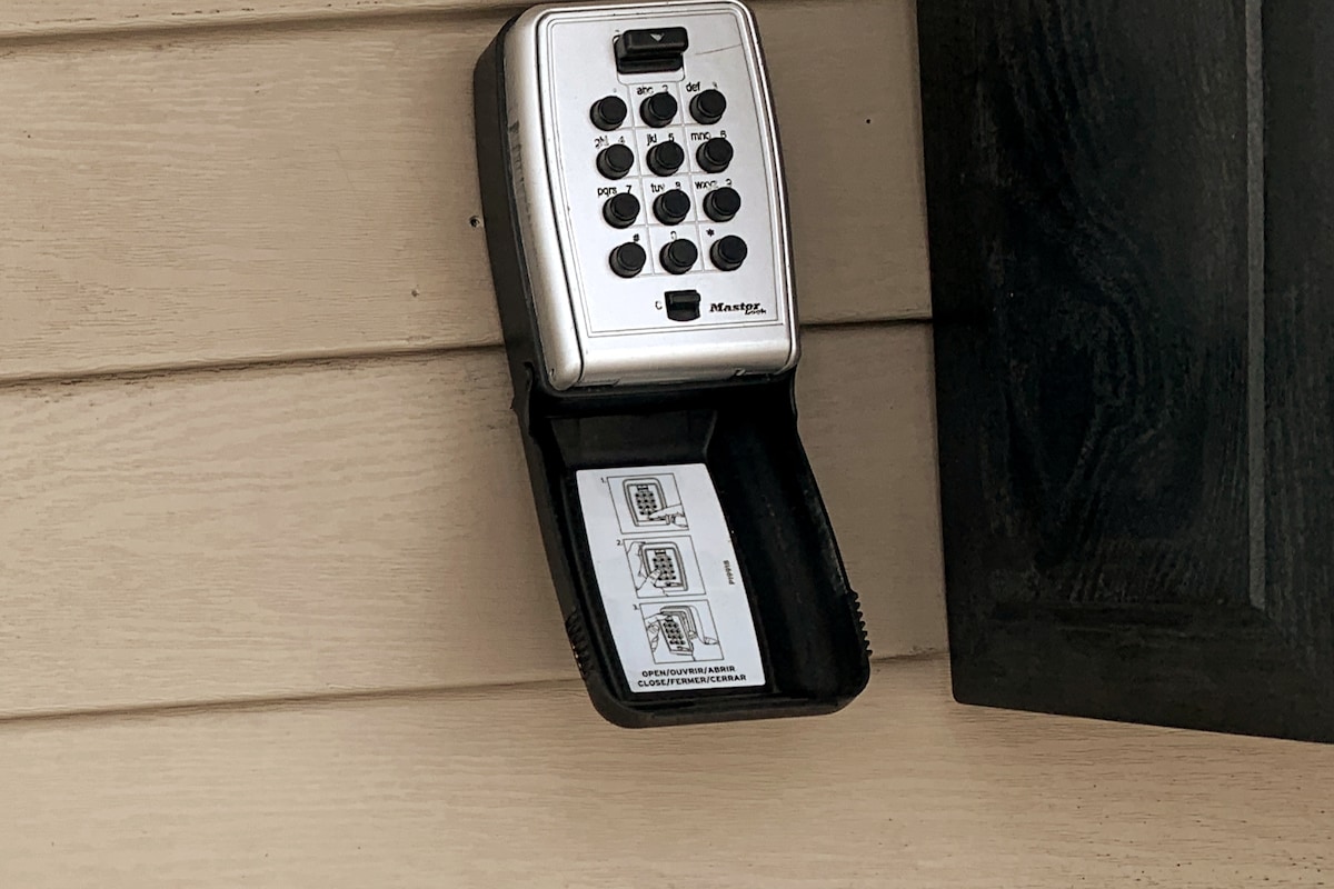 A metal keypad lockbox is mounted on a beige wall. The lockbox is open, displaying a set of instructions inside, with numbers and buttons clearly visible on the keypad. The surrounding area is simple and functional.