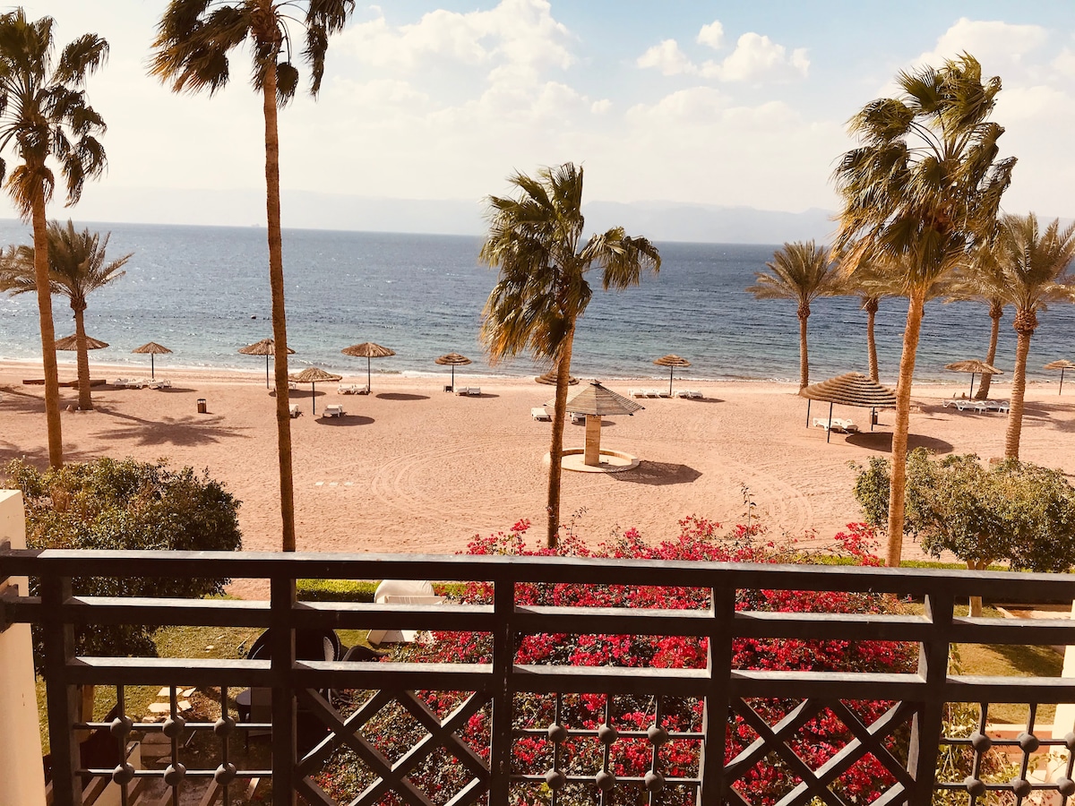 A view of the beach is presented from a balcony, featuring soft sandy shores lined with palm trees and straw umbrellas. The blue sea extends to the horizon, under a partly cloudy sky. Vibrant flowers in the foreground add a touch of color to the scene.