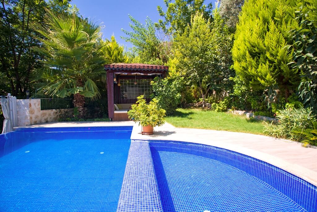 A private pool is framed by lush greenery, featuring vibrant plants and trees. A gazebo is visible in the background, providing shaded seating. The pool is designed with blue tiles, reflecting the sunlight, creating a serene outdoor environment.