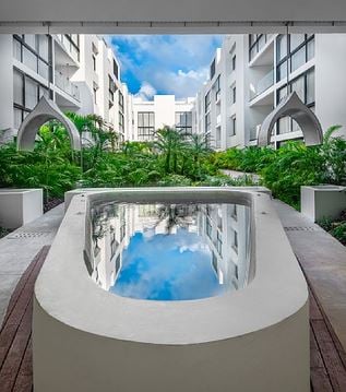 A tranquil courtyard is depicted, featuring a sleek rectangular pool reflecting the surrounding greenery and blue sky. Modern white buildings frame the scene, showcasing the lush vegetation and architectural details that enhance the area's serene ambiance.