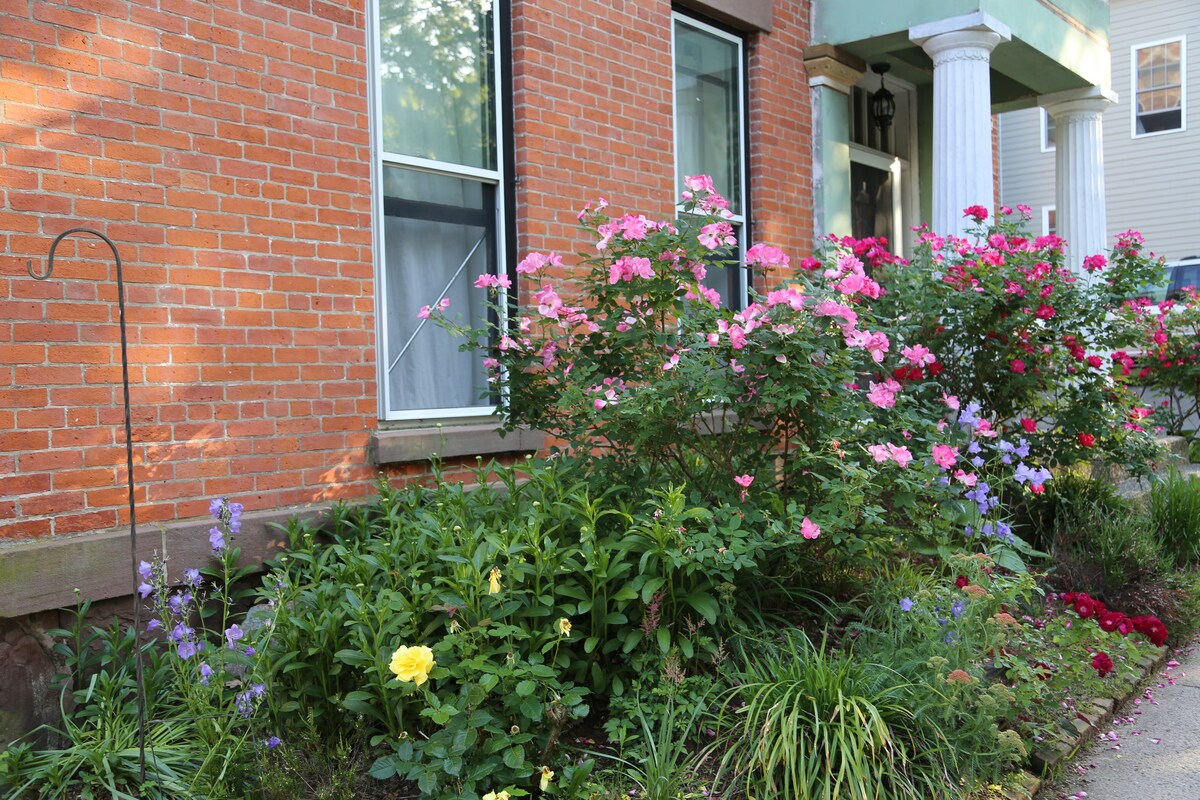 A vibrant garden features blooming pink and yellow flowers in front of a brick home. Lush greenery surrounds the colorful floral displays, with a brick foundation and large windows providing an inviting view of the entrance. Tall white columns are visible on the side of the building.