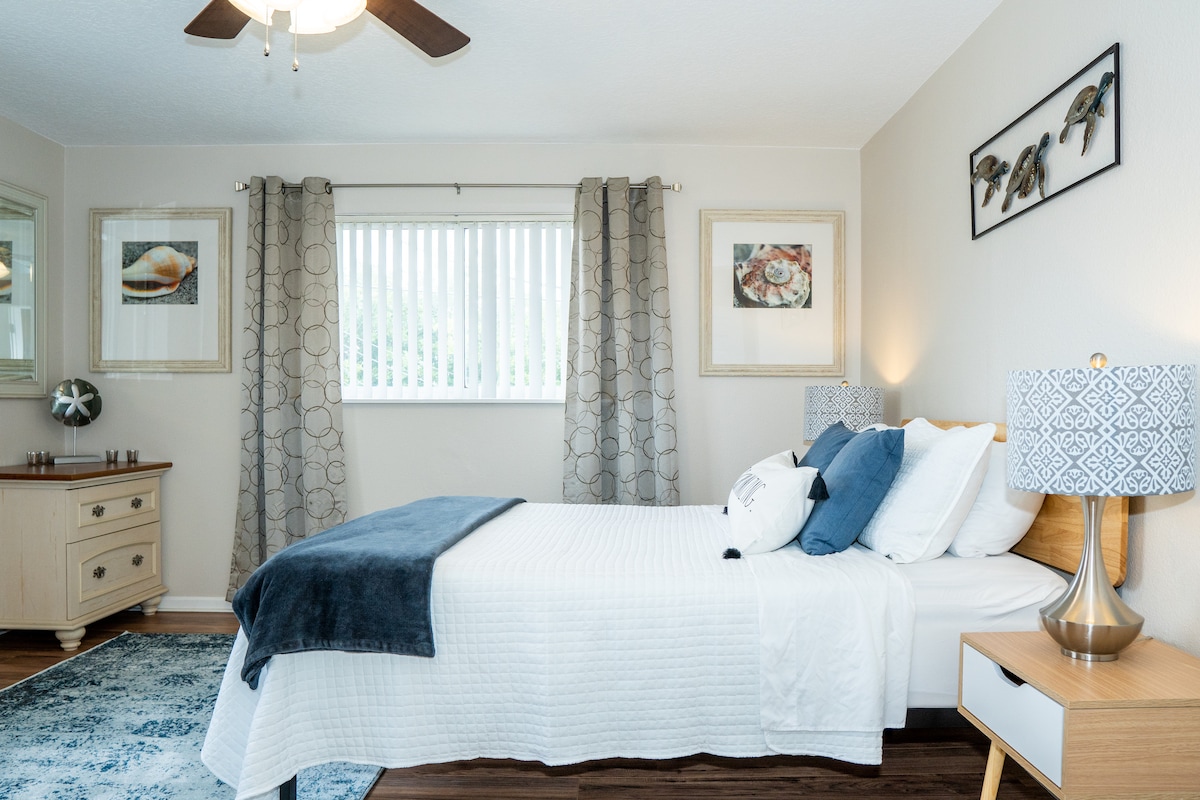 A serene bedroom features a queen-size bed dressed in white linens, with decorative pillows and a navy throw blanket. Soft light filters through vertical blinds, illuminating the space. Artwork adorns the walls, and a dresser with a mirror adds functionality.