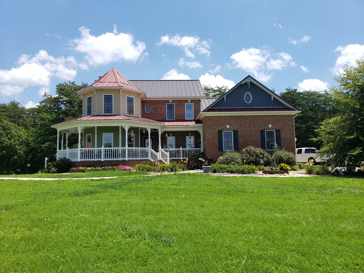 The charming retreat is showcased with a blend of brick and siding, featuring a welcoming front porch and a distinct metal roof. Lush green grass surrounds the home, and colorful flower beds add visual interest, all under a bright blue sky with fluffy clouds.