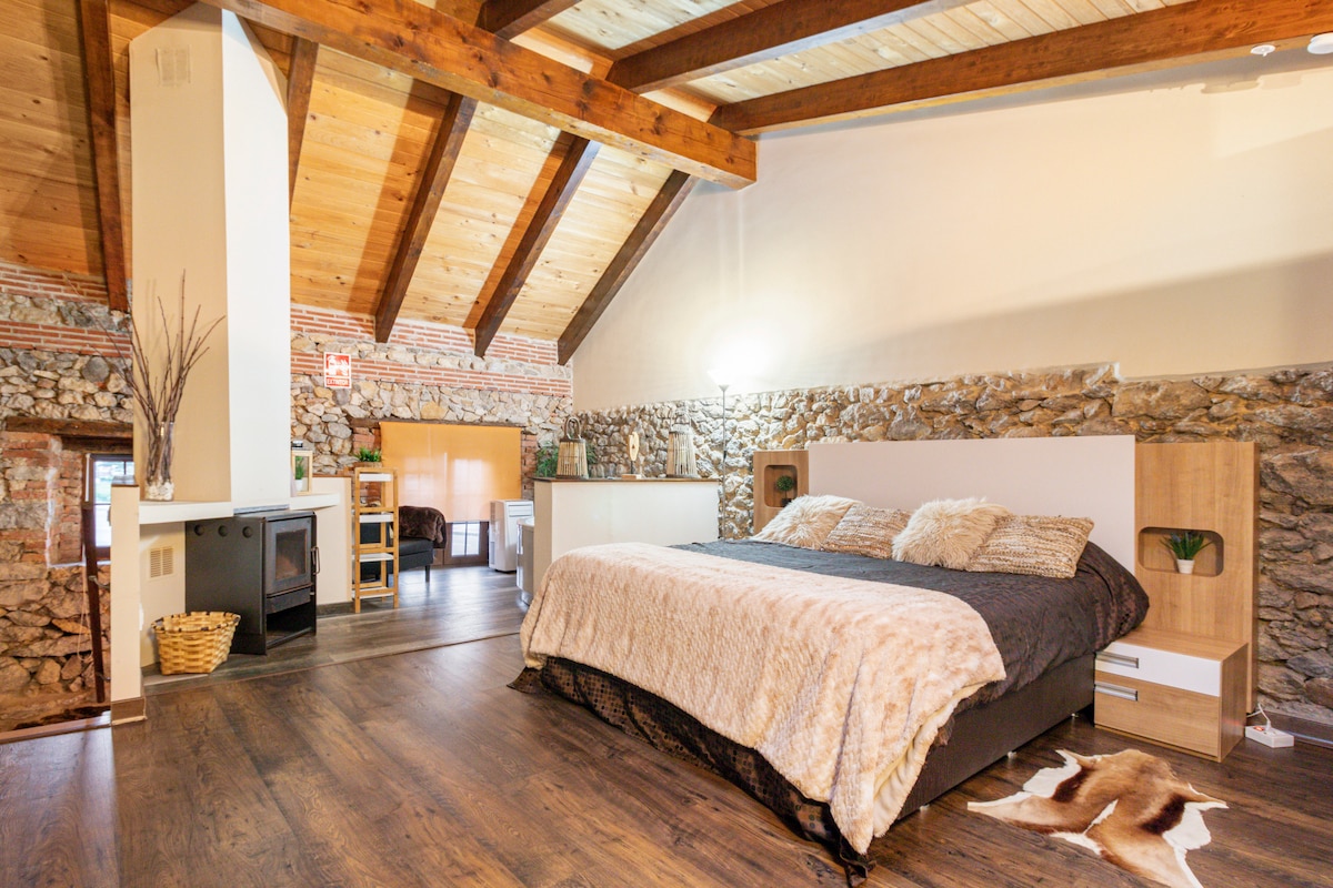 A spacious bedroom features a large bed with stylish bedding, complemented by wooden beams and stone walls. A cozy fireplace is positioned nearby, adding a charming touch. Natural light illuminates the room, creating a warm and inviting atmosphere.