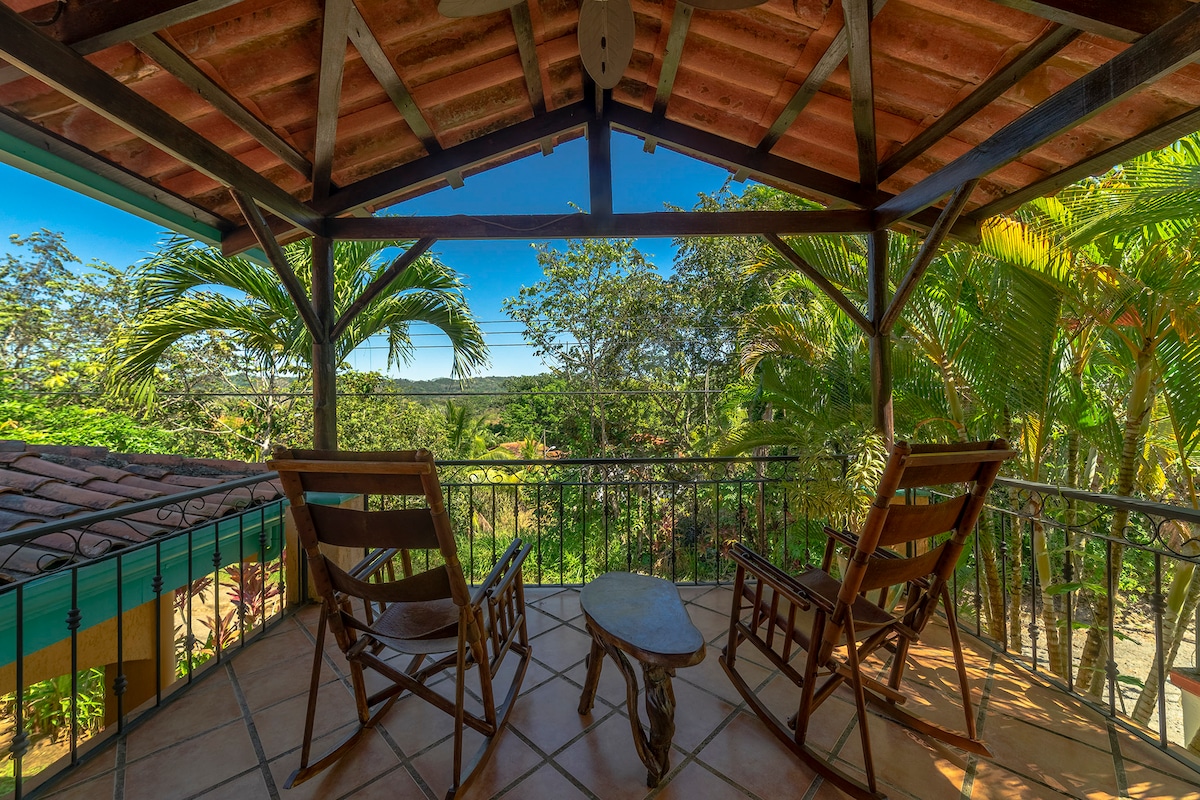 A scenic outdoor gazebo is furnished with two wooden rocking chairs and a small table. Lush greenery surrounds the area, with trees and tropical plants visible. The roof features wooden beams, and the tiled floor adds a natural touch to the inviting space.