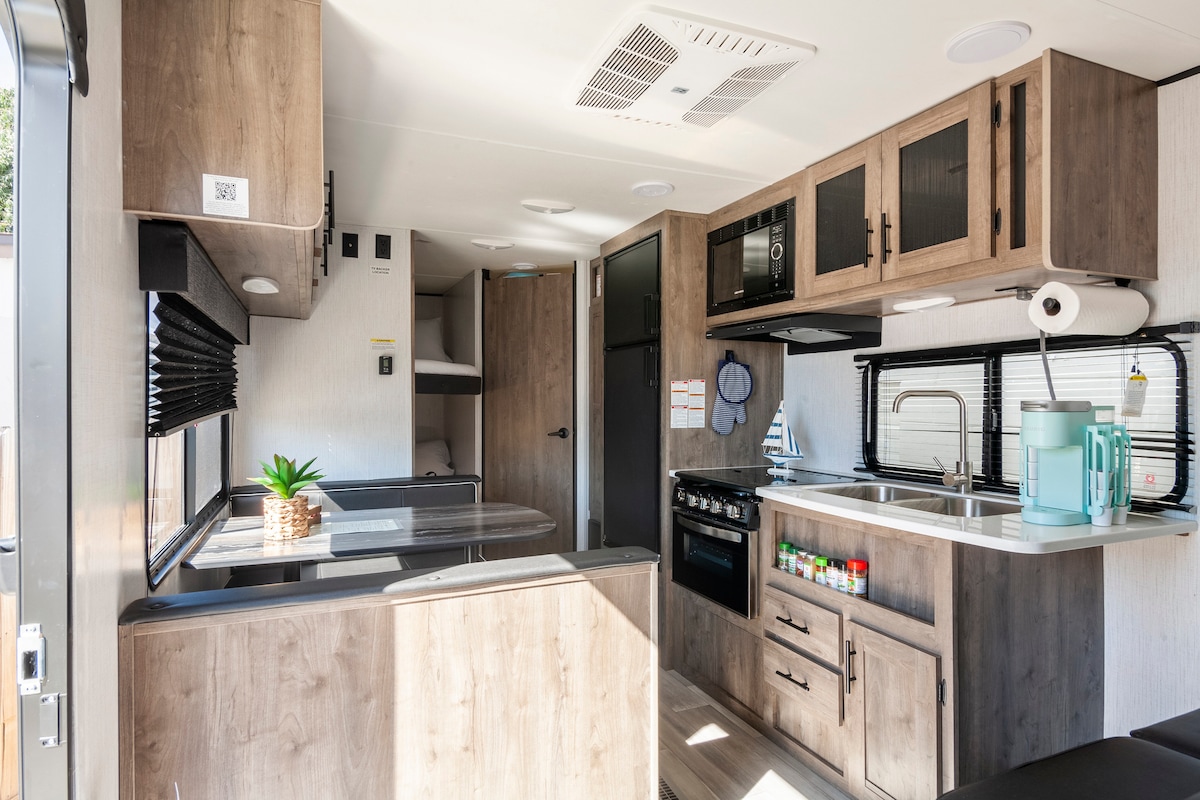 A fully equipped kitchen area is showcased with wooden cabinetry and modern appliances. A dining table with seating is visible, along with a stove and refrigerator. Light enters through a nearby window, creating a bright and functional space for meal preparation.