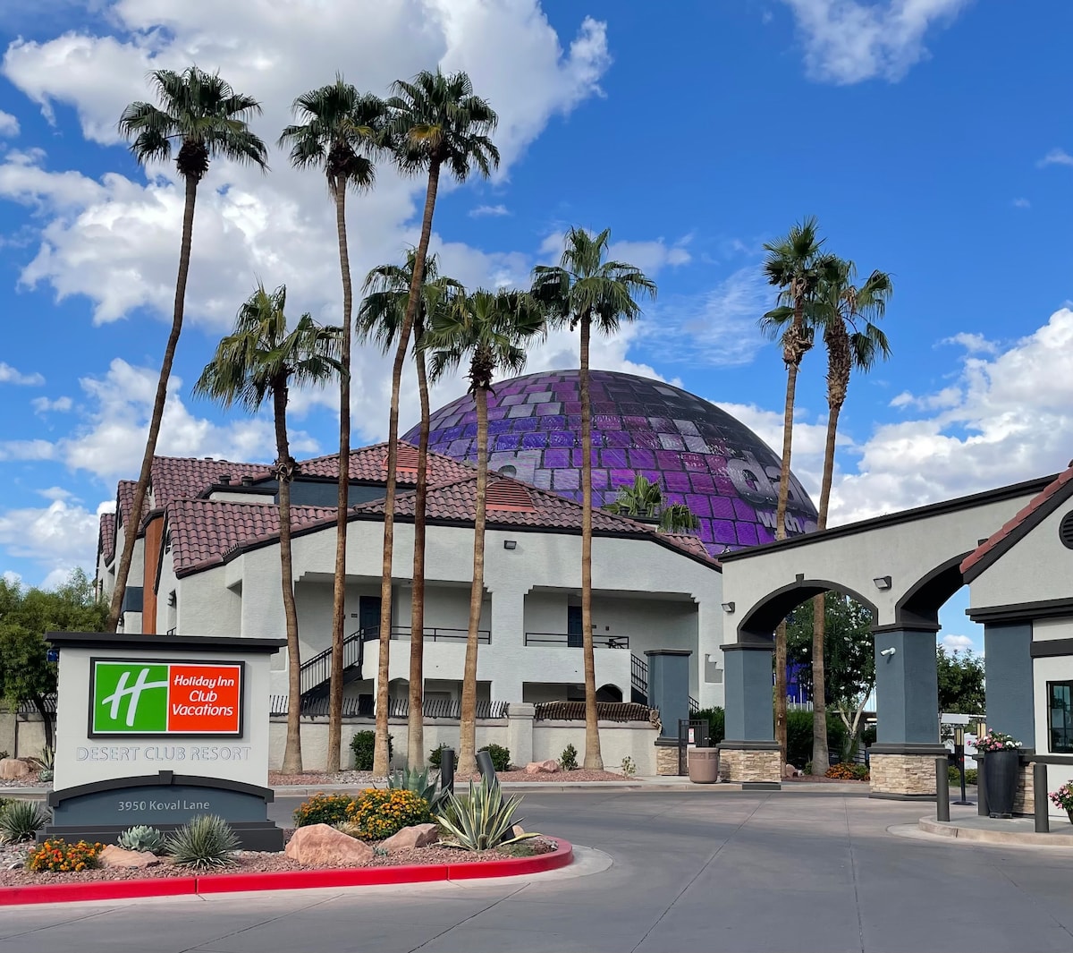 The entrance to the Holiday Inn Desert Club Resort is highlighted by a well-maintained sign and surrounded by palm trees. A striking dome structure with purple accents is visible in the background, set against a clear blue sky with scattered clouds.