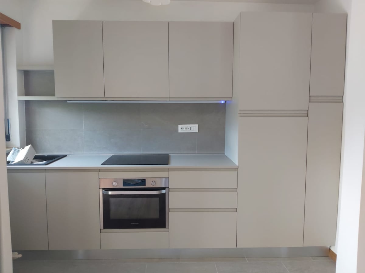 A modern kitchen is displayed, featuring sleek gray cabinetry with multiple storage options. An integrated oven and cooktop are positioned beneath a subtle countertop. The design emphasizes functionality with clean lines and minimalistic fixtures, illuminated by soft overhead lighting.
