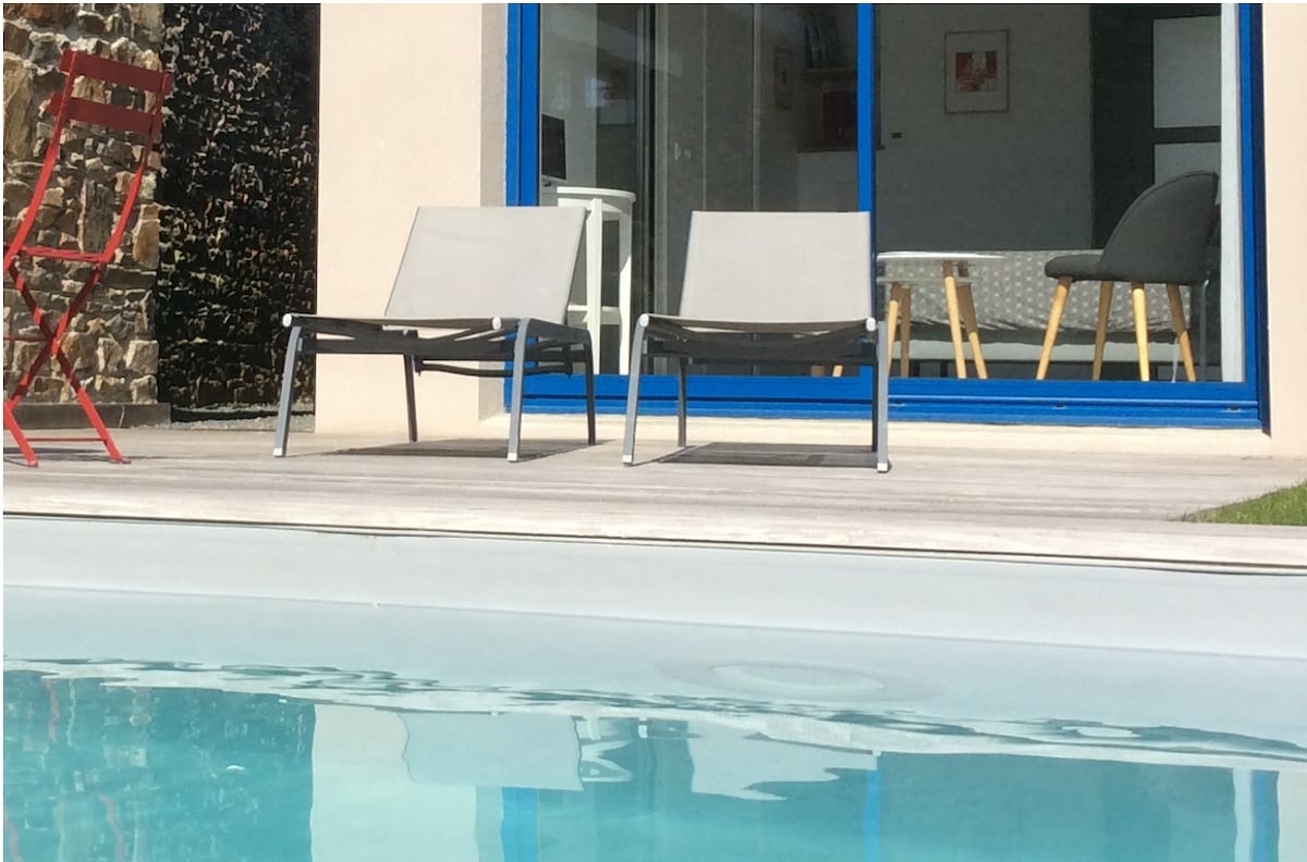 Two gray lounge chairs are positioned by the edge of a heated swimming pool. Light reflects off the water's surface. A bright blue frame surrounds large glass doors that lead to an interior space, enhancing the outdoor connection.