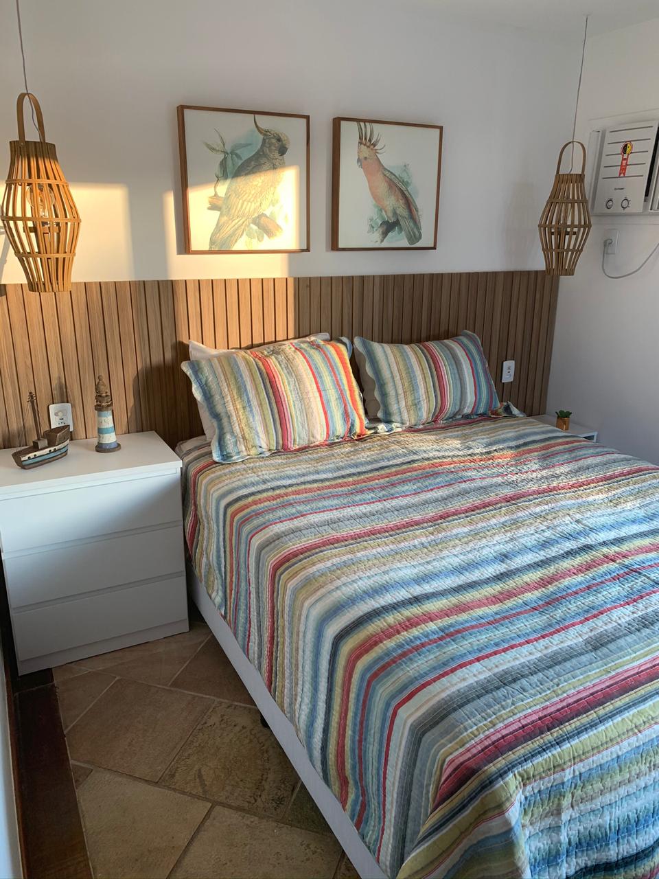 A cozy bedroom features a spacious bed adorned with a striped blanket and matching pillows. Two hanging light fixtures provide soft illumination, while decorative artwork depicting birds adorns the wall. A nightstand with a decorative item sits beside the bed, enhancing the room's charm.