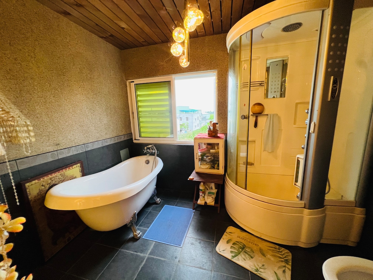 A spacious bathroom features a freestanding bathtub and a glass-enclosed shower. Natural light enters through a window with green slats, illuminating the room's textured walls. Decorative light fixtures hang from the ceiling, and a bath mat is placed on the floor.