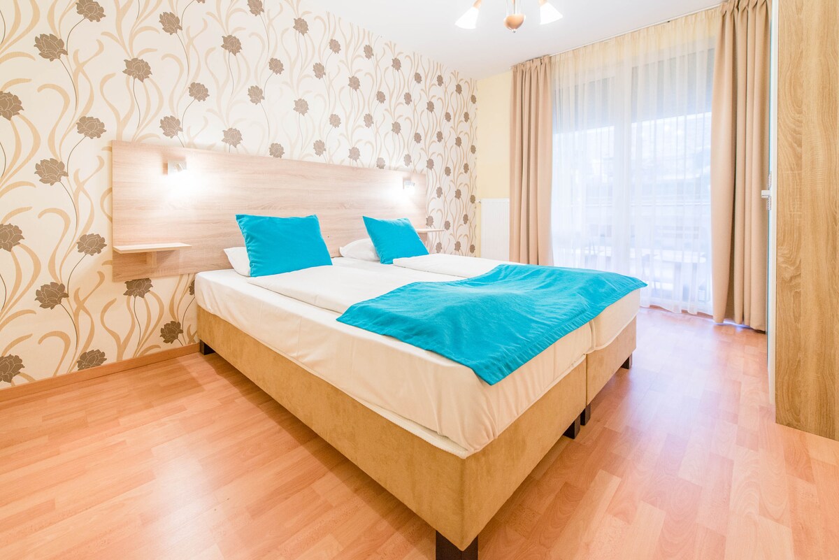 A spacious sleeping area features two beds with turquoise blankets, complemented by a light wood headboard. The walls are adorned with a subtle floral design, and natural light enters through the large window covered with sheer curtains.
