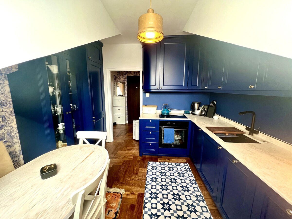 A compact kitchen features deep blue cabinetry and a marble countertop. A round dining table sits nearby, surrounded by white chairs. A patterned rug lies on the wooden floor, complementing the space. Bright light is provided by a pendant lamp hanging above.
