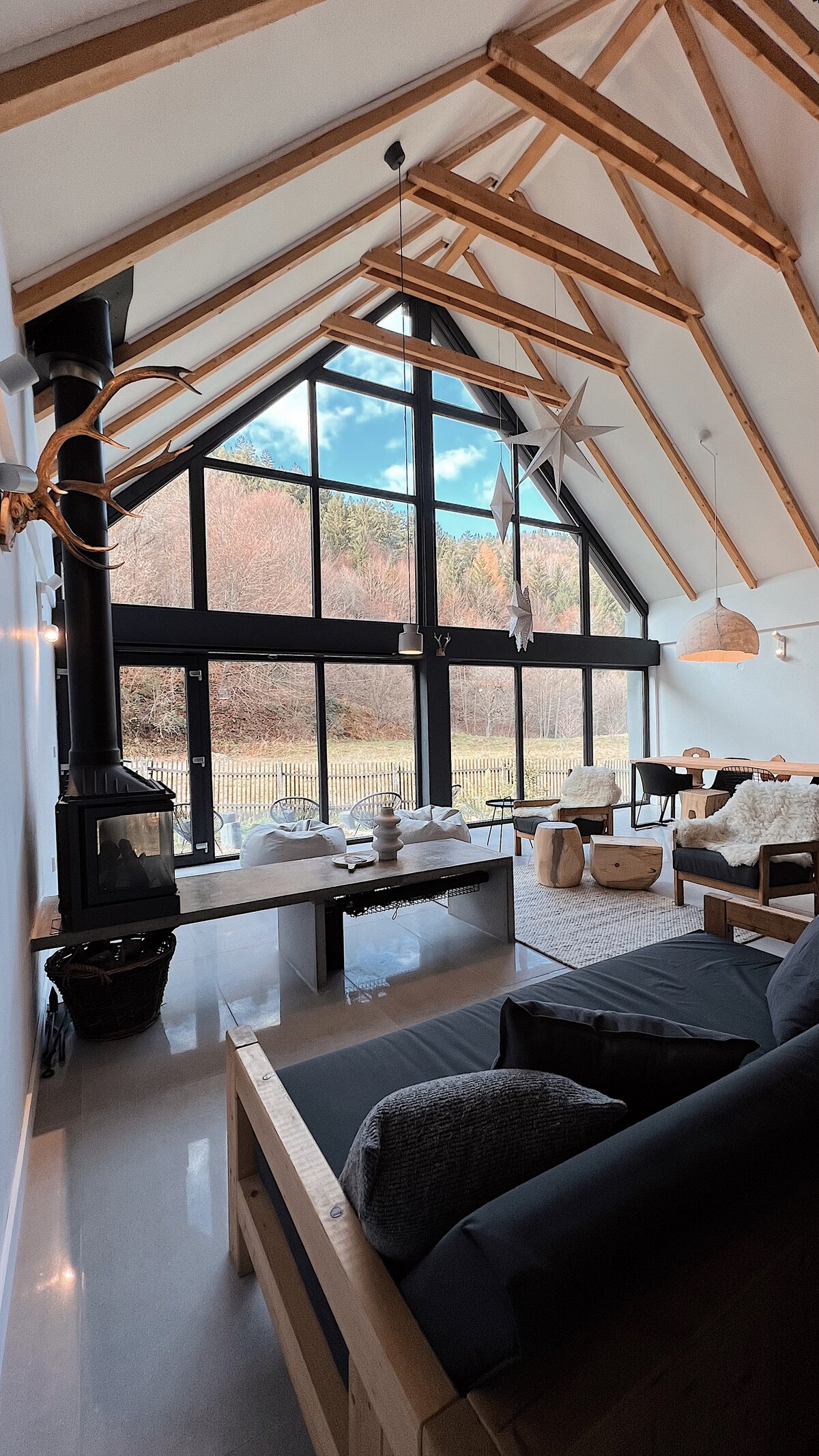 The open living area is defined by tall, slanted ceilings and large glass windows that invite natural light. A modern fireplace is present, complemented by wooden furnishings and soft textures. The view of nature outside presents a serene backdrop to the cozy interior.