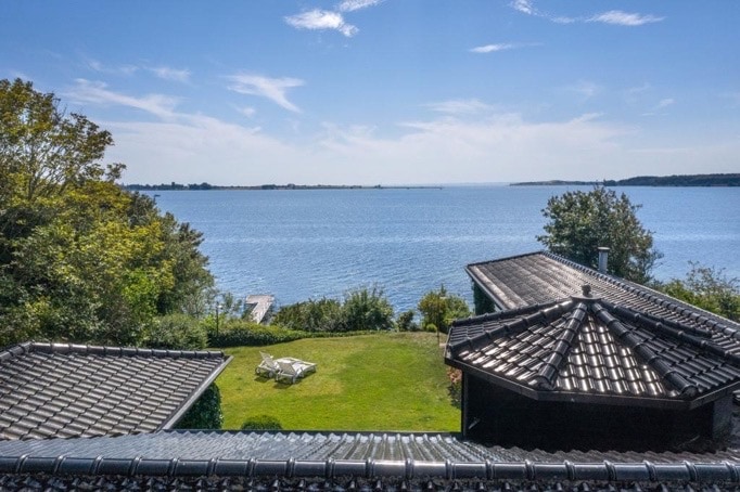 An expansive view of the water is captured, with a bright blue sky above. Lush green gardens are visible in the foreground, offering a tranquil space for relaxation. The image includes rooftops of nearby houses, providing context to the serene, waterfront setting.
