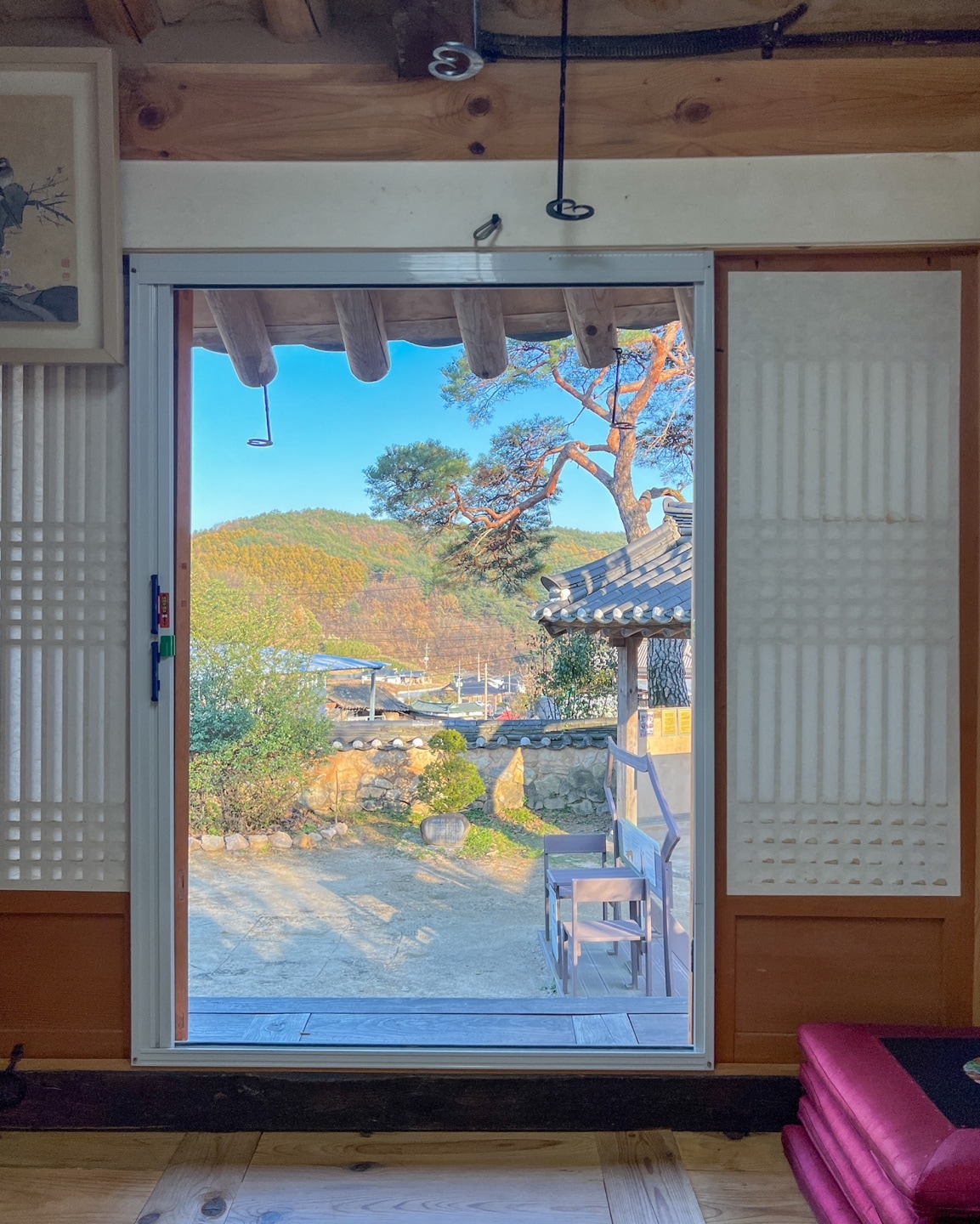 The image captures a doorway framed by traditional sliding panels, opening to a view of lush hills and a serene outdoor landscape. Sunlight filters through, illuminating the interior space, while the nearby pine trees add a touch of nature to the picturesque scene.