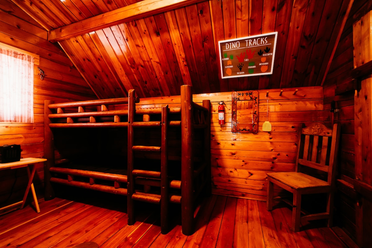 The interior of the rustic cabin features warm wooden walls and ceiling beams. A pair of sturdy bunk beds is positioned against the wall, complemented by a wooden chair and table nearby. Natural light filters through a small window with sheer curtains.