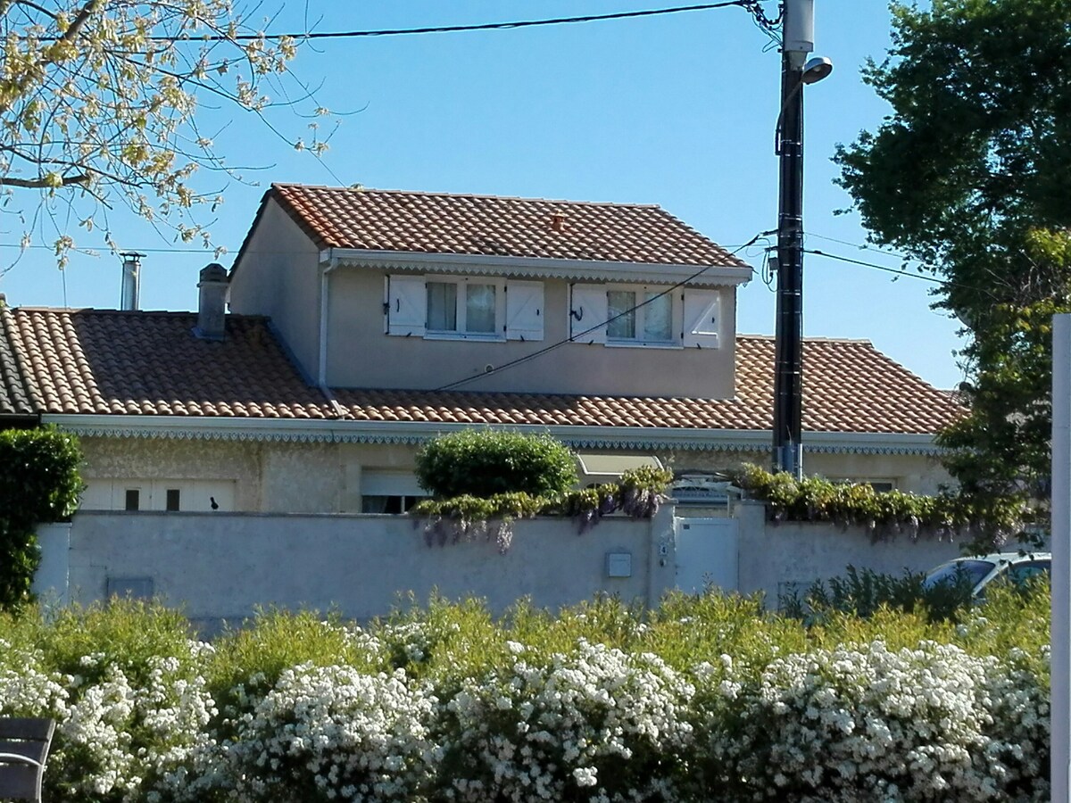 A two-story house is surrounded by well-maintained greenery and flowering shrubs. The entrance features a simple white door, framed by a small garden. The roof is covered with traditional red tiles, and the windows are adorned with shutters, situated under a clear blue sky.