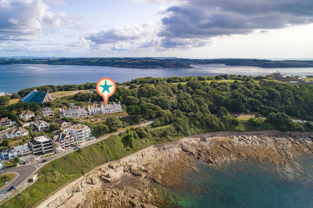 An aerial view captures the coastal landscape, featuring a collection of contemporary buildings set against lush greenery. The shimmering waters of Falmouth Bay reflect the sky, with a distant shoreline hinting at the historical Pendennis Castle.