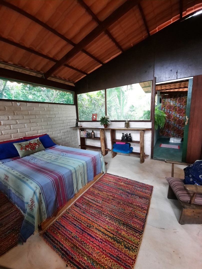 A cozy sleeping area is equipped with a spacious bed adorned with a colorful striped blanket. Natural light filters through nearby windows, illuminating the earthy tones of the room. A wooden shelf displays decorative items, and a vibrant rug adds warmth to the floor.