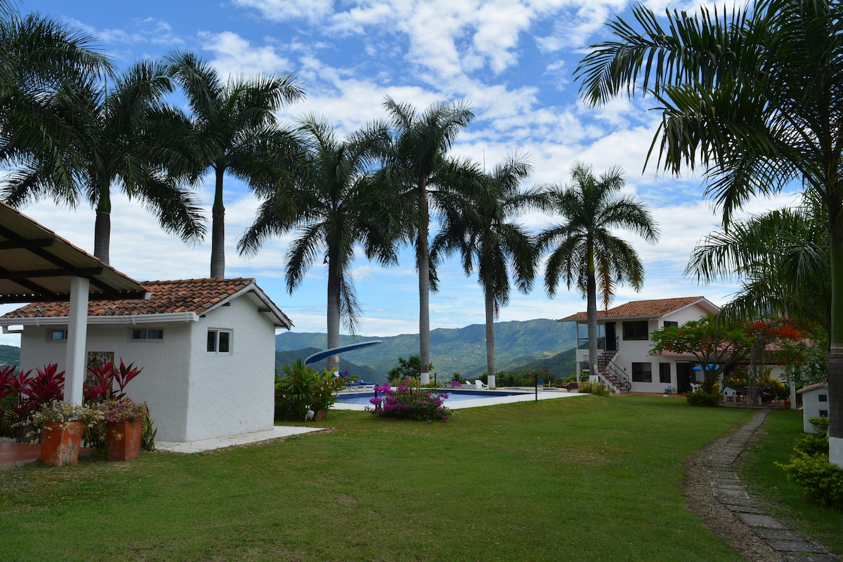A scenic outdoor area featuring palm trees and well-maintained grass. Two structures are visible: a white building with a terracotta roof and another larger structure in the background. The expansive pool reflects the bright sky and is surrounded by various plants.