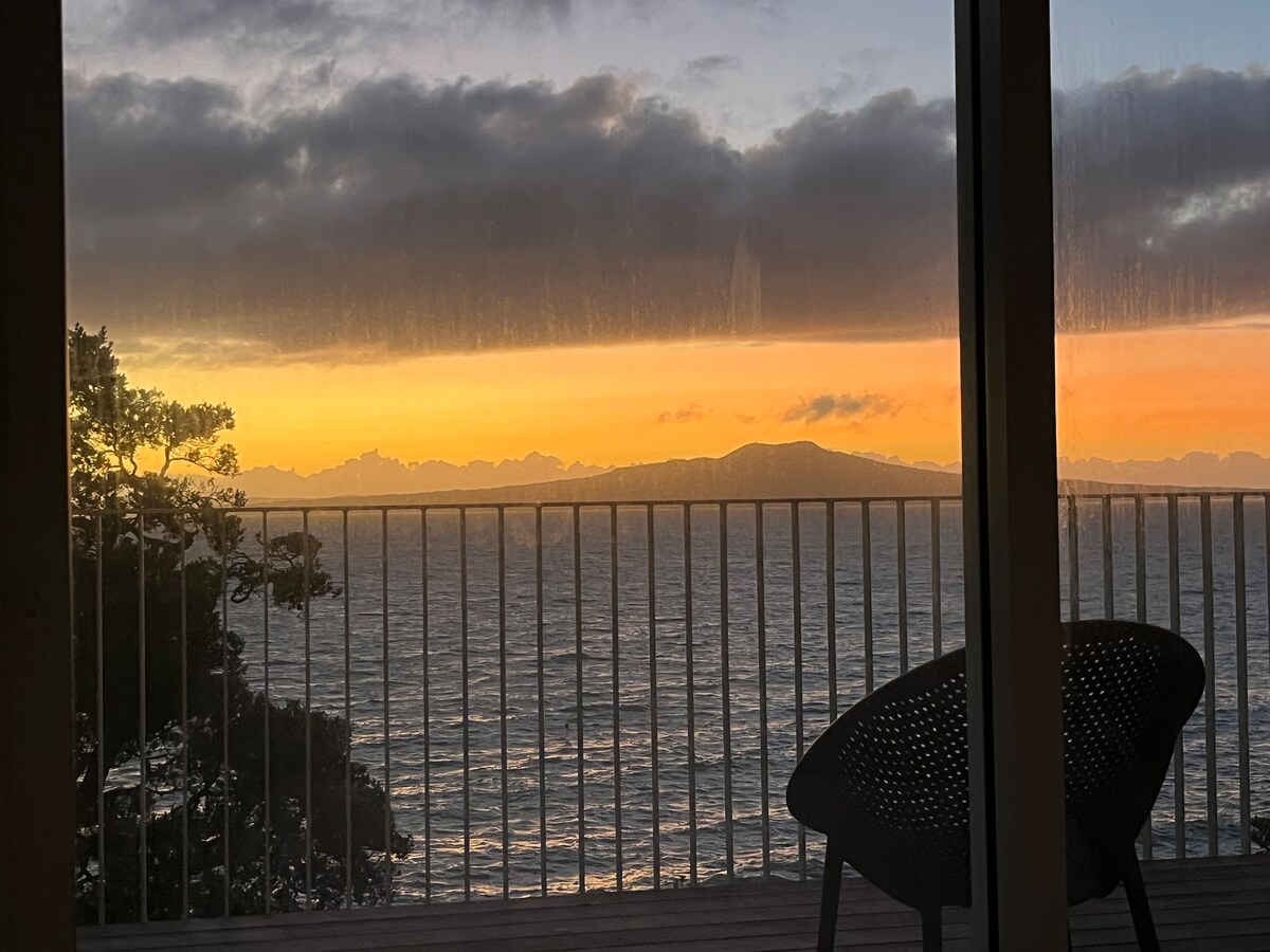 A stunning view is captured from the apartment's sliding doors, showcasing a vibrant sunrise over the ocean. Rangitoto Island is visible in the distance, while the soft light reflects on the water's surface. A black outdoor chair is partially seen in the foreground.