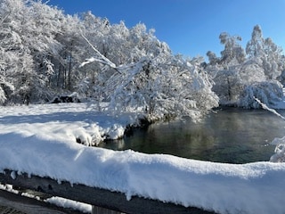 A snow-covered landscape is displayed, featuring a serene body of water surrounded by frosted trees. A wooden railing is partially visible at the foreground, with a thick layer of snow resting on top. The clear blue sky enhances the tranquil winter scene.