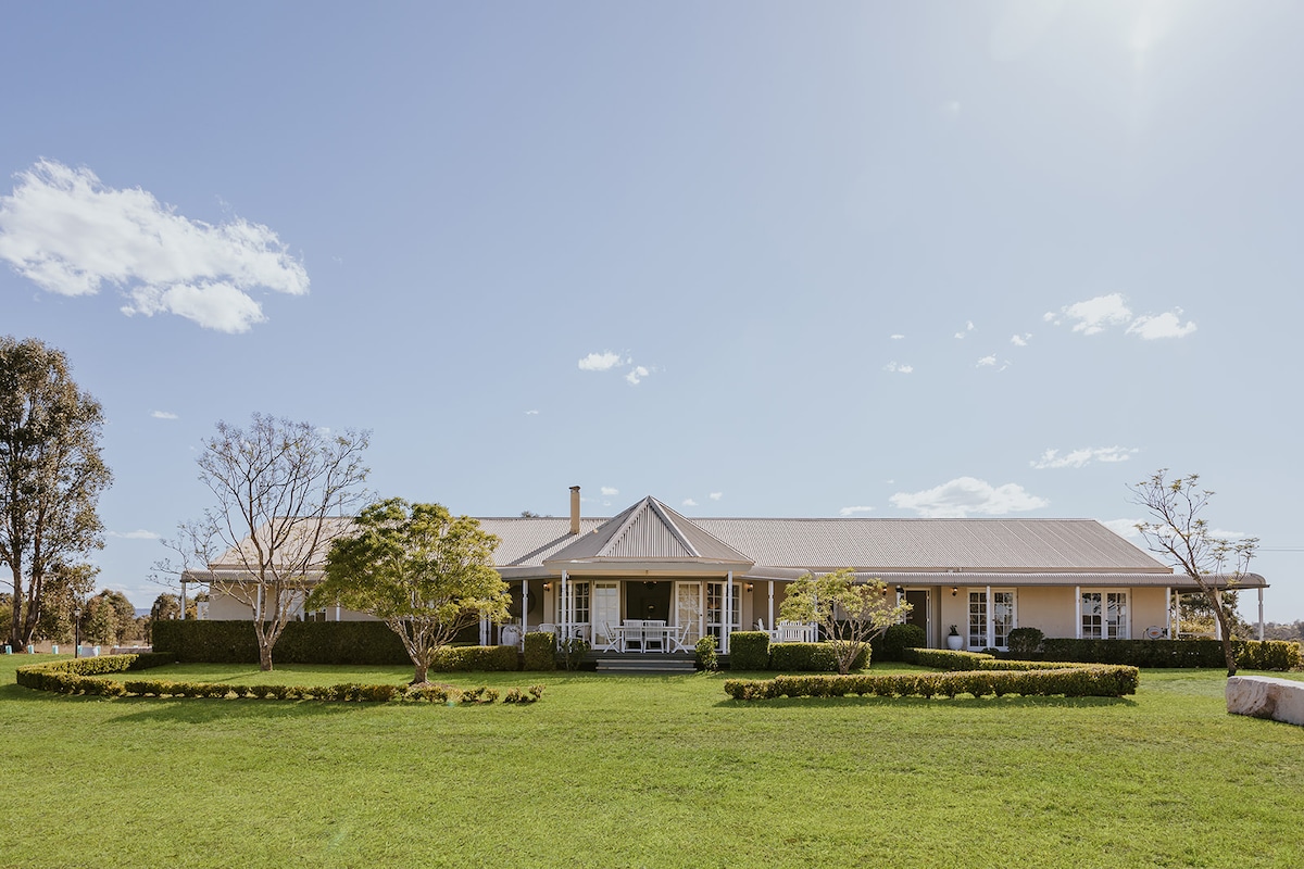 A colonial-style house is set against a clear blue sky, featuring a large wrap-around porch. The expansive lawn is bordered by manicured shrubs and trees, creating an inviting outdoor space that complements the property's tranquil rural setting.