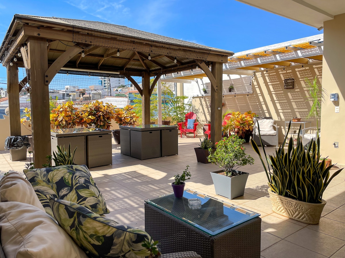 A common terrace area features a wooden gazebo surrounded by potted plants and seating arrangements. Comfortable sofa seating is visible, along with a coffee table. Sunlight filters through the canopy, creating a relaxed outdoor atmosphere.