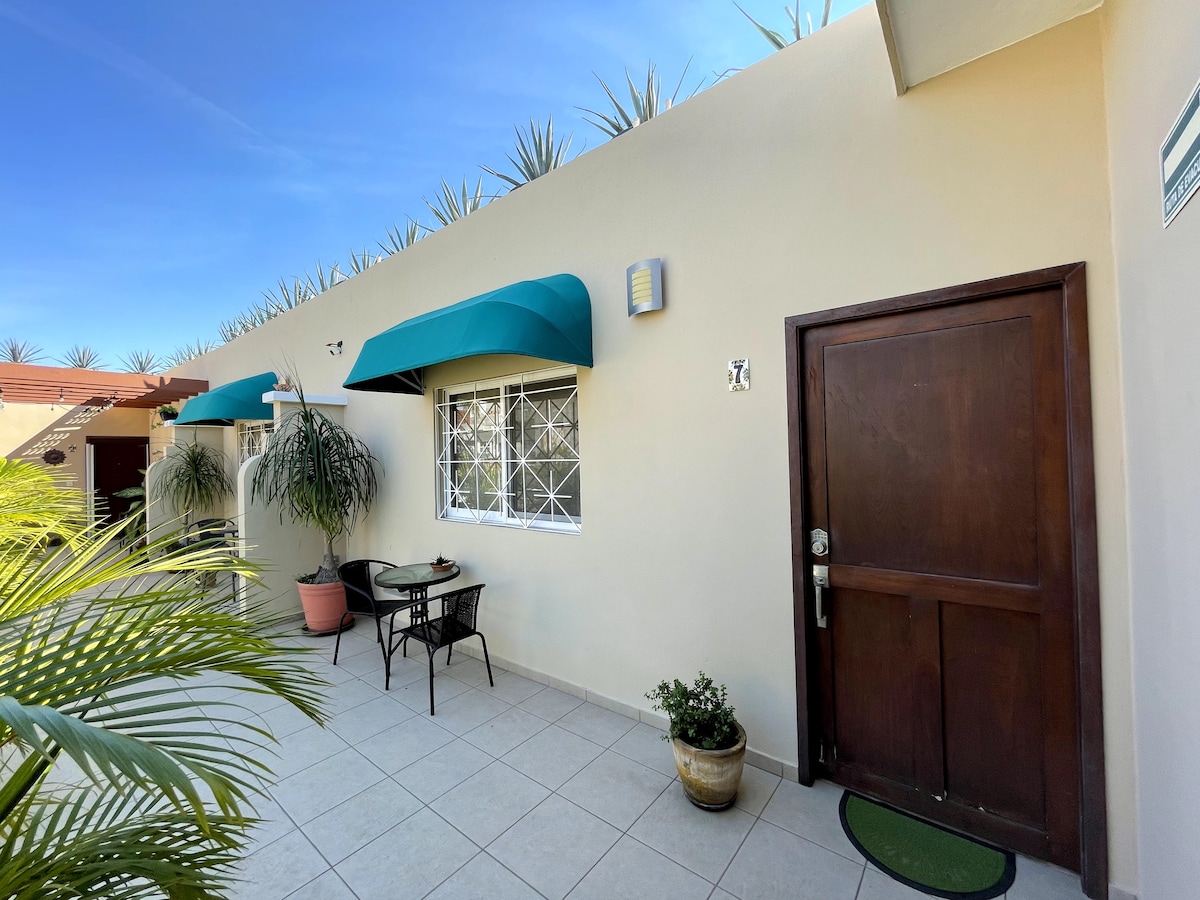 A welcoming entrance is presented, featuring a wooden door and a small patio area. A bistro table with two chairs is positioned beside the entrance, surrounded by potted plants. Green awnings provide shade, and tiles create a smooth pathway.