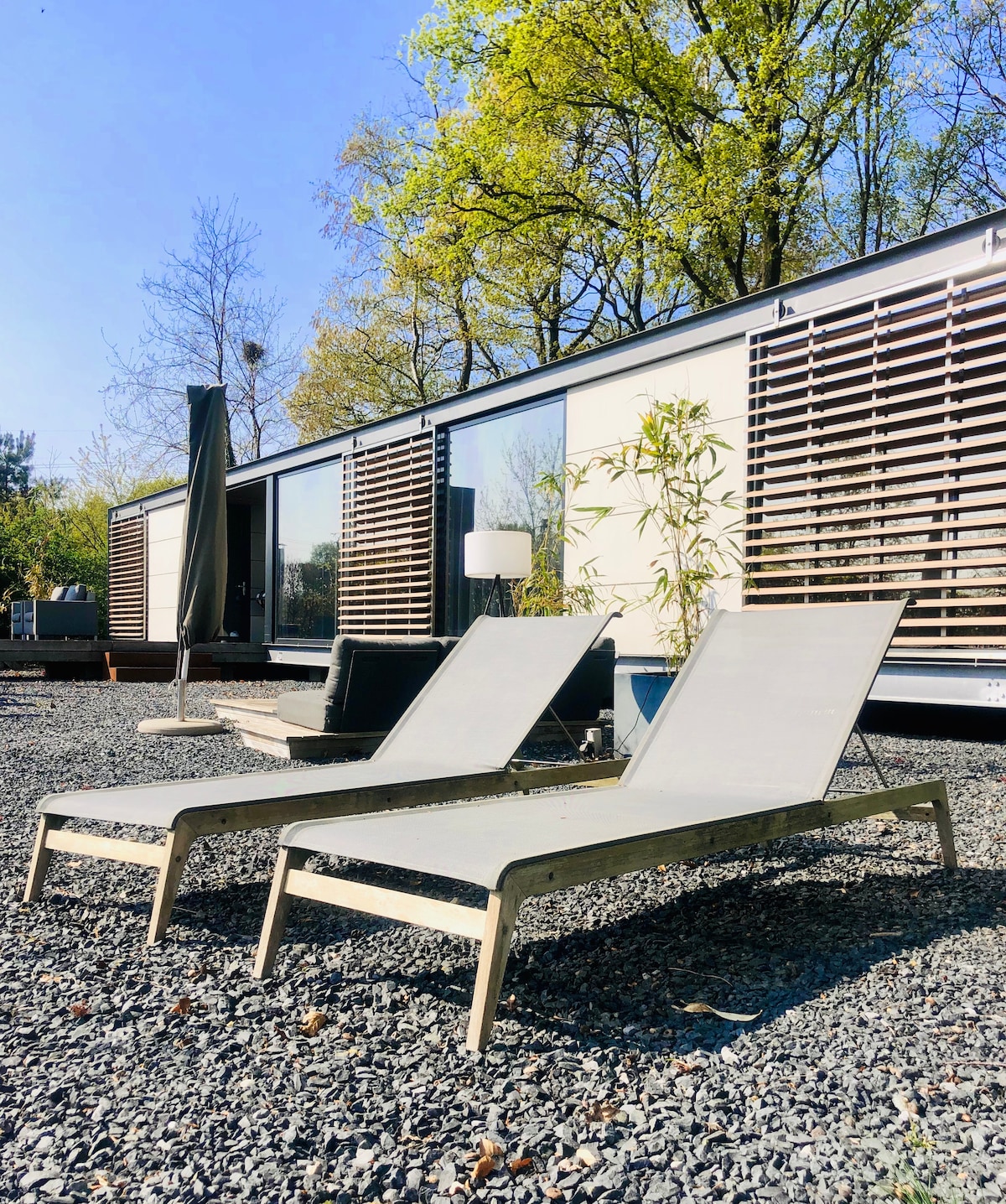 Two lounge chairs are positioned on a gravel surface, with a backdrop of a modern structure featuring large glass windows and wooden slatted shades. Lush greenery surrounds the area, enhancing outdoor relaxation.