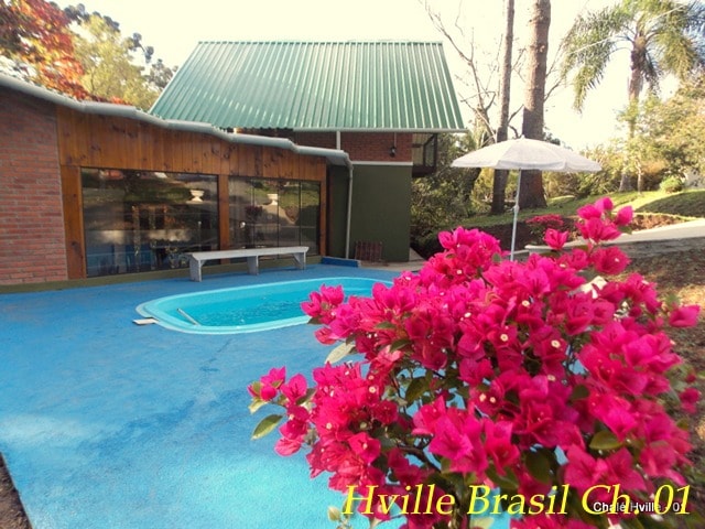 The outdoor area features a small pool surrounded by vibrant flowers and a shaded umbrella. A bench is positioned nearby, complemented by a backdrop of greenery and a house with a sloped roof, enhancing the serene outdoor environment.