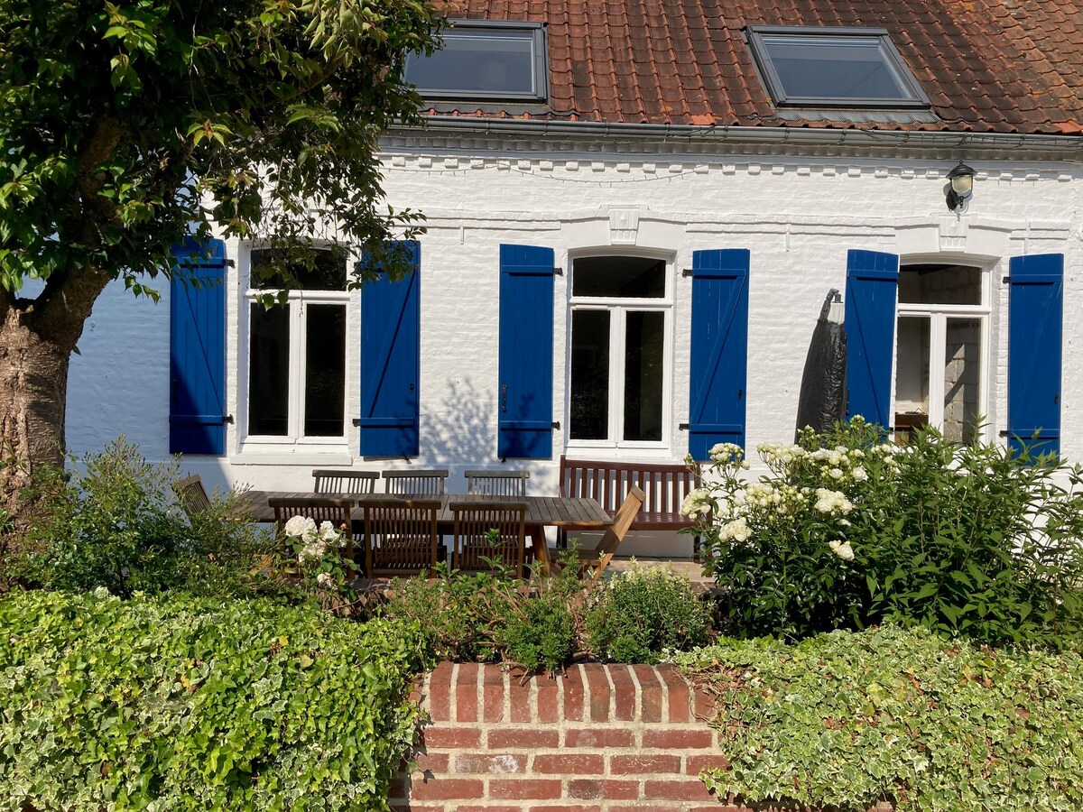 The exterior of the farmhouse displays charming white walls with blue shutters framing the windows. A wooden table and chairs are positioned in a landscaped area, surrounded by greenery and flowering plants, creating a serene outdoor space.