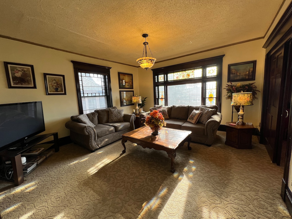 A spacious living room features large stained glass windows allowing natural light to illuminate the area. Comfortable sofas are arranged around a wooden coffee table, accented by a floral arrangement. A flat-screen TV is positioned in the corner, complementing the warm decor.