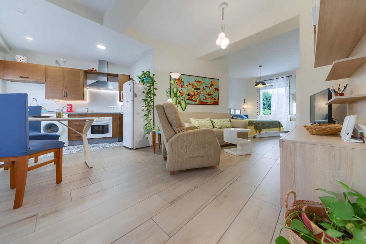 An open living area features a comfortable seating arrangement, complemented by a colorful wall art piece. A modern kitchen is seen in the background, equipped with essential appliances. Large windows allow natural light to fill the space, and greenery adds a touch of nature.