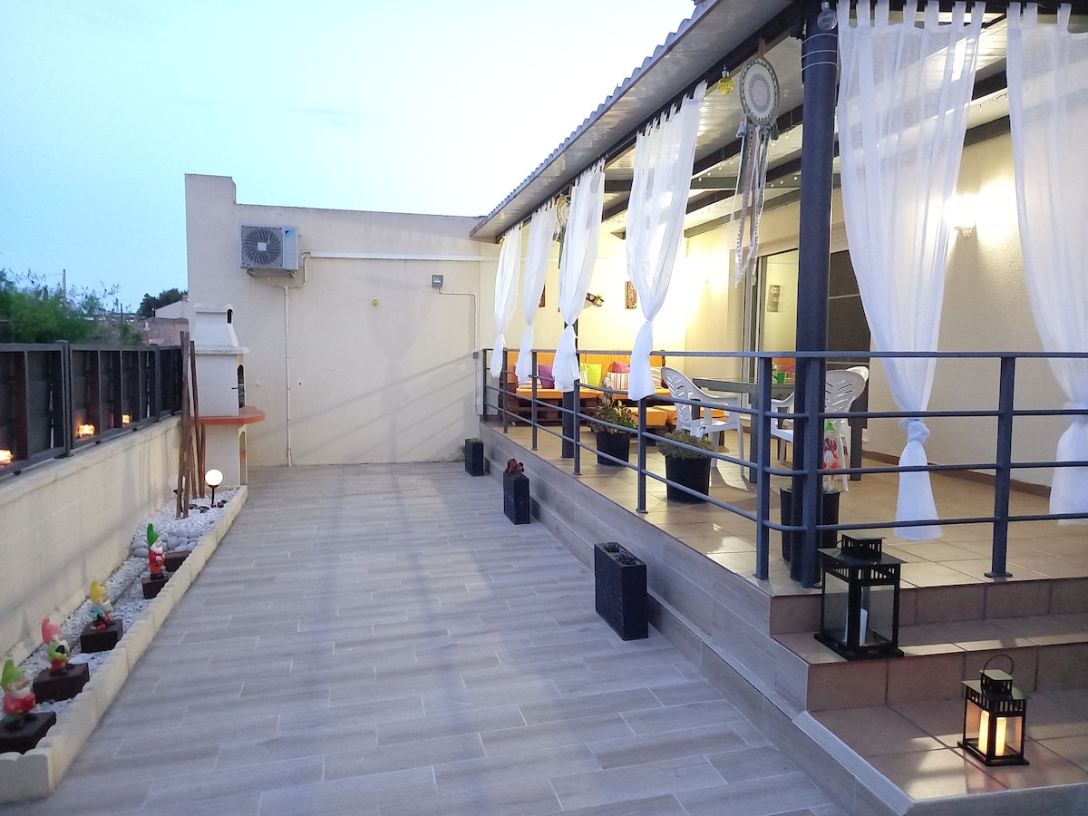 A spacious terrace is visible, featuring a tiled floor and draped white curtains creating a soft ambiance. Comfortable seating areas are arranged under a covered roof, with potted plants and lanterns adding subtle decoration. The evening sky casts a gentle light over the scene.