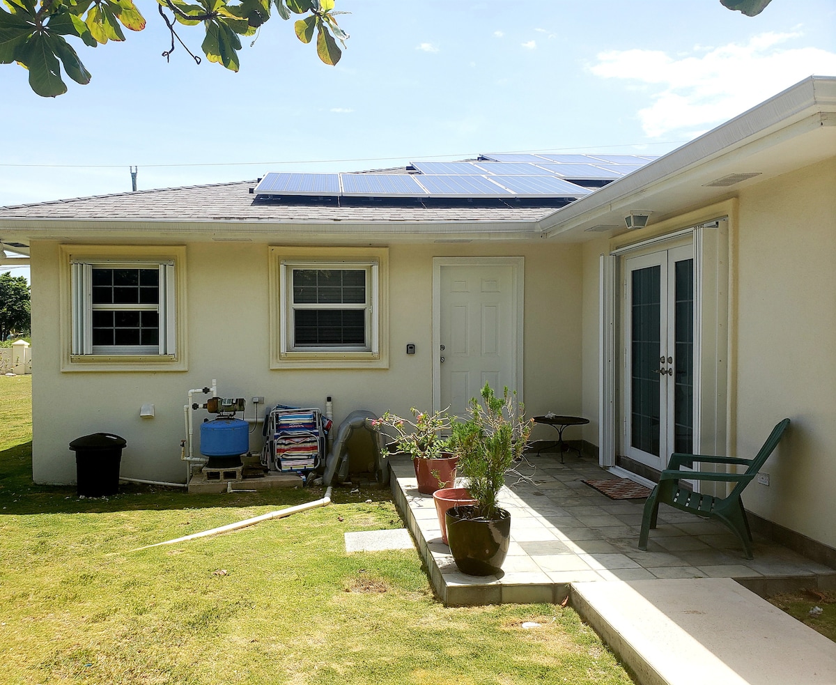 The exterior view of the apartment showcases a light-colored structure with solar panels on the roof. A small patio area is visible, featuring a green chair and potted plants. Nearby, laundry and utility items are neatly arranged, with a grassy yard providing a spacious outdoor environment.