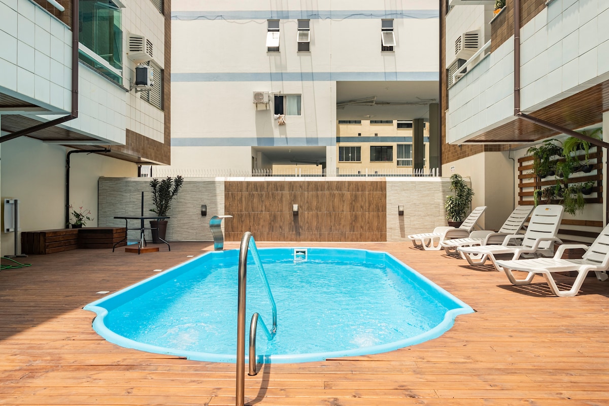 A rectangular swimming pool is framed by wooden decking and six deck chairs. Cool blue water reflects sunlight, while a small ladder offers access to the pool. Surrounding greenery and modern air conditioning units are visible on nearby walls.