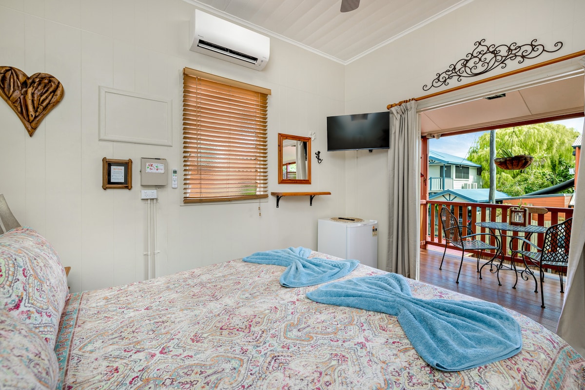 A spacious sleeping area features a queen bed adorned with a patterned quilt and two rolled towels. Natural light is welcomed through the nearby French doors, leading to a balcony furnished with a table and chairs. A small fridge and a wall-mounted TV are included.
