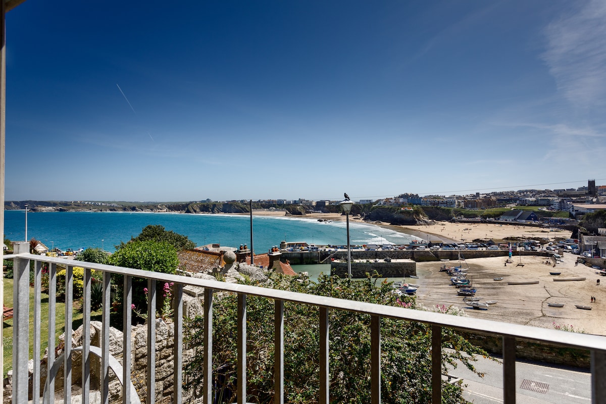 A clear view of Newquay Harbour and its coastline is captured from the Juliet balcony. The vibrant blue sea contrasts with the sandy beach and bustling harbor area below. The scene is framed by the balcony's white railing, providing a picturesque outlook.