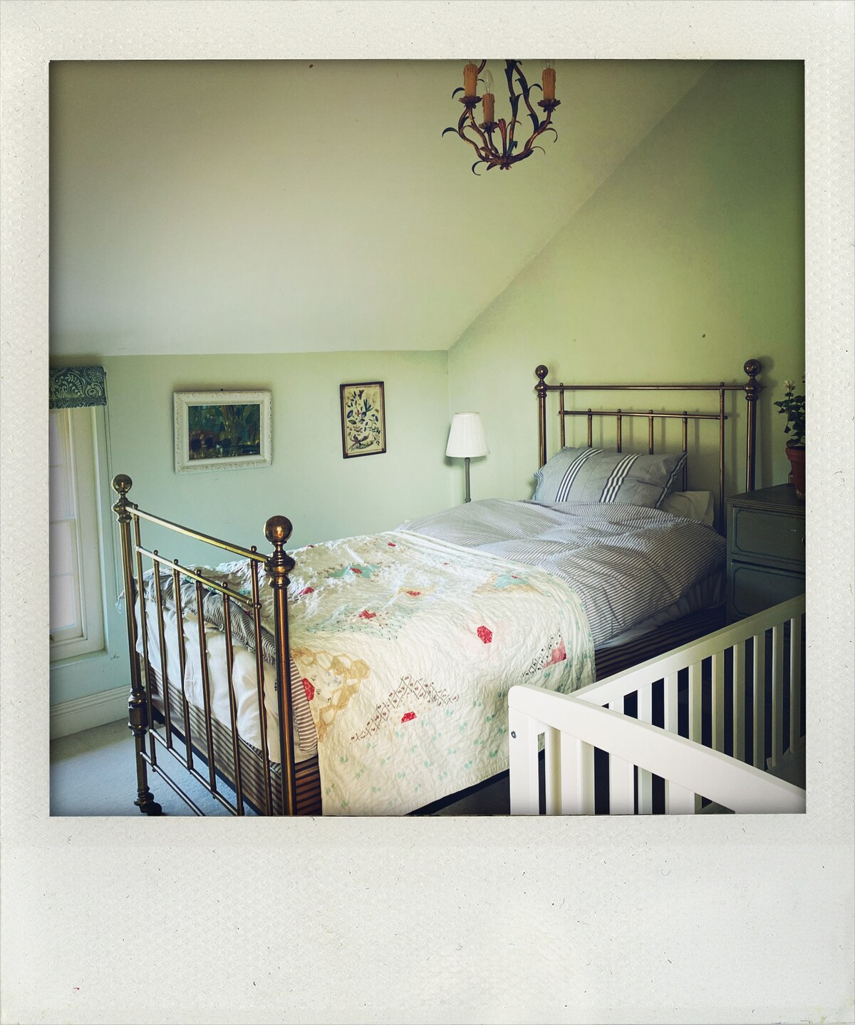 A cozy bedroom features a metal bed frame with a floral patterned quilt. Soft green walls create a calming atmosphere, while a bedside lamp and framed artwork add decor. Natural light enters through a window, enhanced by a sheer curtain. A wooden railing provides separation.