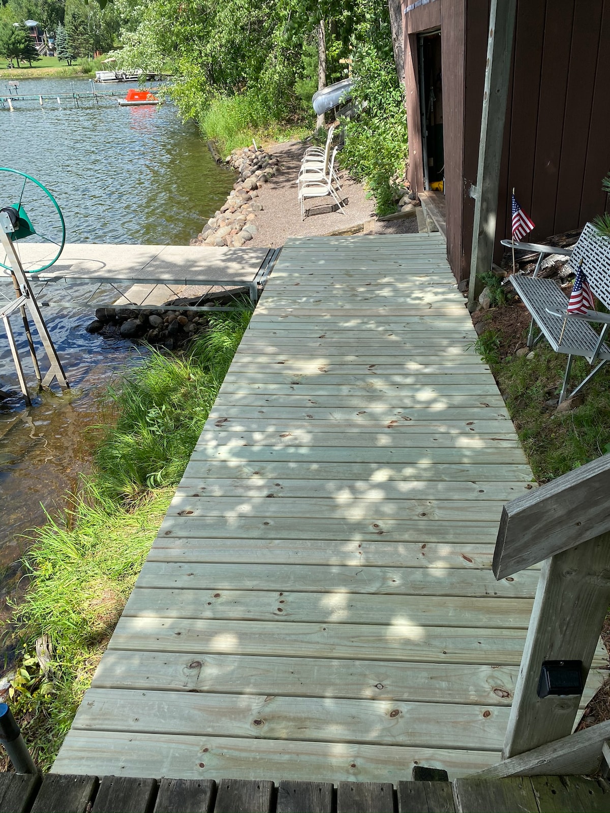 A wooden walkway leads from the cabin towards the water, flanked by green grass and dotted with small rocks. A few chairs are positioned nearby, and small flags are visible, adding to the lakeside environment. The calm water reflects shadows from surrounding trees.