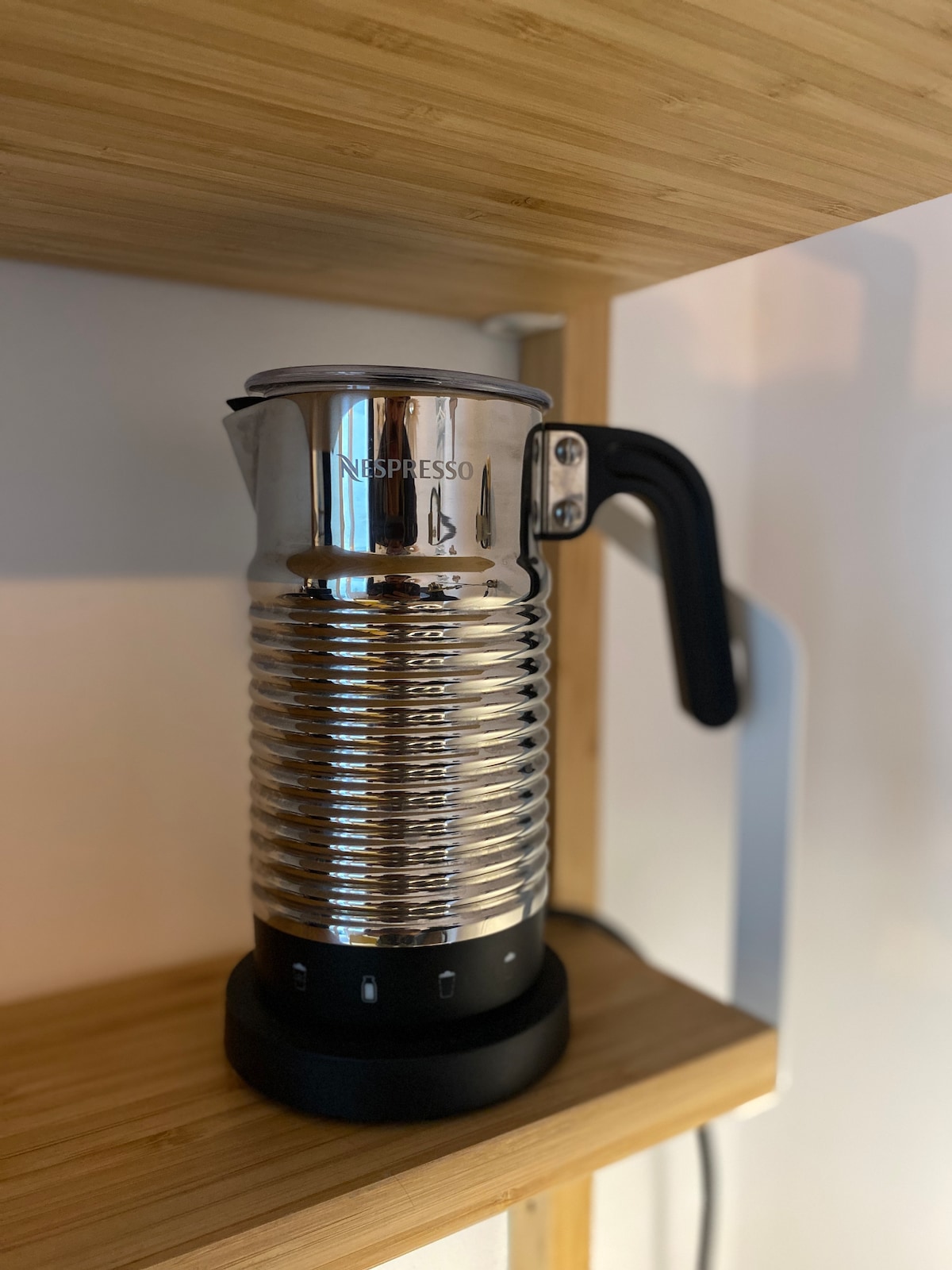 A sleek Nespresso coffee maker is positioned on a wooden shelf. The stainless steel exterior features a spiral design, and the base includes control buttons for ease of use. The surrounding cabinet offers a minimalist and modern aesthetic.