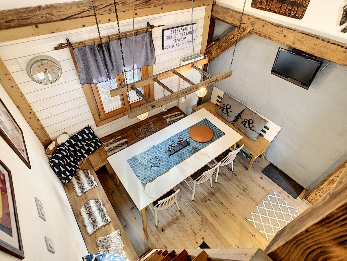 An overhead view captures a bright open plan dining area featuring a long table set for meals. Cozy seating with decorative cushions is arranged around the table, and a small TV is mounted on the wall, positioned above a wooden bench adorned with decorative pillows.